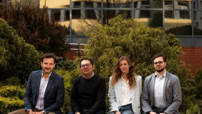 Cztery osoby siedzące na ławce na tle zieleni i budynku